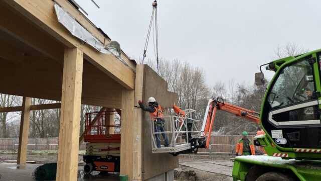 Crane installation of a Dun Agro prefabricated timber-framed hempcrete wall panel, serving as a monolithic, carbon-negative building envelope.